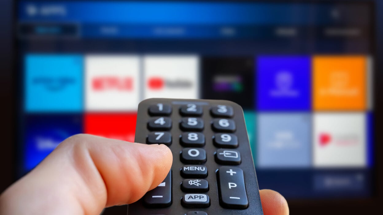 a right hand holds a remote control pointed at a TV with streaming app icons shown on the screen