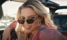 A blonde teen girl smiles from a car wearing sunglasses.