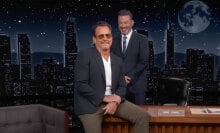A man in sunglasses sits on a talk show desk while another man in a suit stands behind laughing.