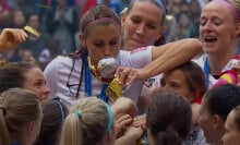 A group of women in soccer shirts hold a trophy aloft while one of them kisses it.