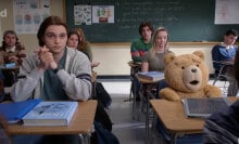 A CGI teddy bear sits at a high school desk in a classroom with other students.
