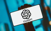A hand holding up a phone displaying the black and white ChatGPT logo.