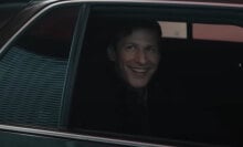 A man sitting in the back seat of a black car with the window down stares out grinning.