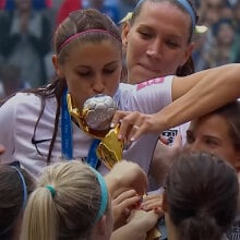 A group of women in soccer shirts hold a trophy aloft while one of them kisses it.