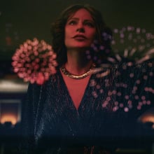A stylish woman stands leaning on a wall as fireworks explode in the background.