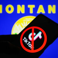 A crossed-out TikTok logo is seen on a smartphone in front of the flag of the state of Montana on a PC monitor.
