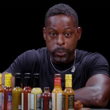 A man sits in front of a table of different sauce bottles, blowing out his cheeks with his eyes wide.