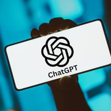 A hand holding up a phone displaying the black and white ChatGPT logo.