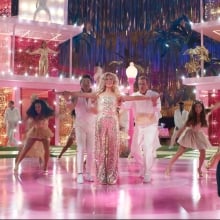 A group of coordinated Barbies and Kens dance on a brightly lit, pink dance floor. A woman in a black shirt interprets in ASL in the bottom right corner of the screen.
