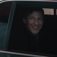 A man sitting in the back seat of a black car with the window down stares out grinning.