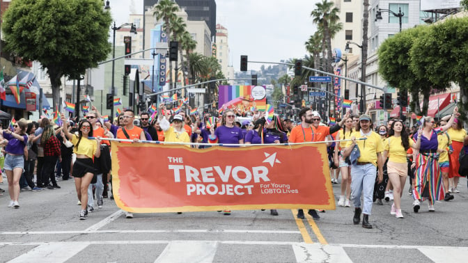 A group of people march down a street during a Pride parade. They are holding a large orange Trevor Project banner. 