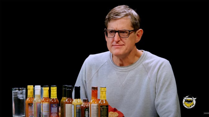 A man with glasses sits at a table covered with bottles of sauce.