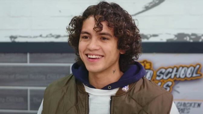 A man with longish curly hair sits at a table in a chicken shop, grinning.