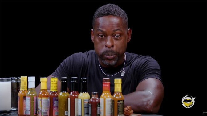 A man sits in front of a table of different sauce bottles, blowing out his cheeks with his eyes wide.