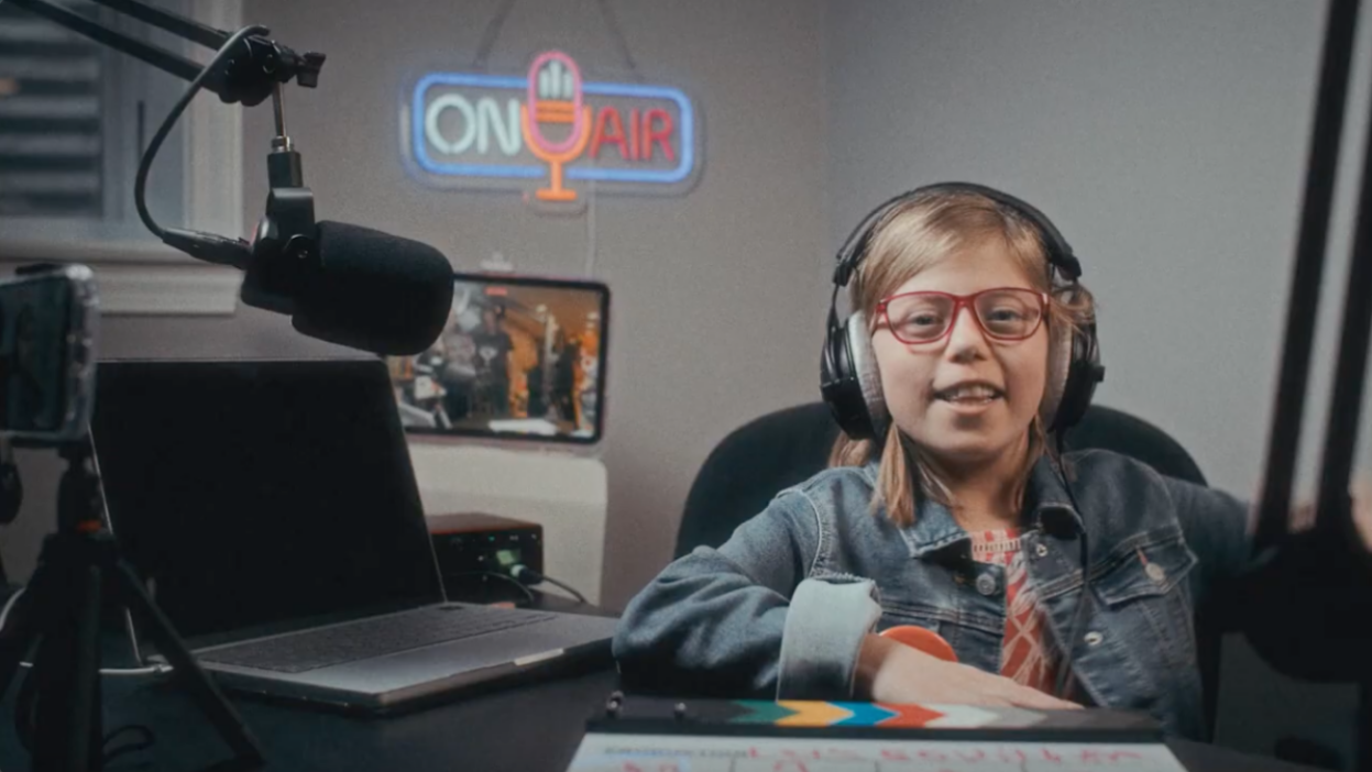 A young girl sits in front of a radio microphone wearing headphones. An "on air" sign glows behind her. 