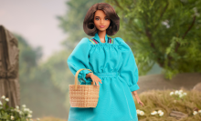 A barbie with short brown hair stands in a long turquoise dress holding a woven basket.