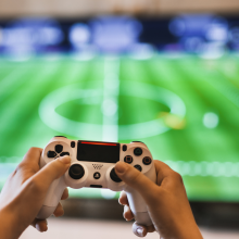 Hands holding a gaming controller in front of a television displaying a sporting event.