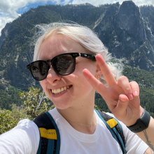 woman wearing sunglasses holding up a peace sign with mountains in the background