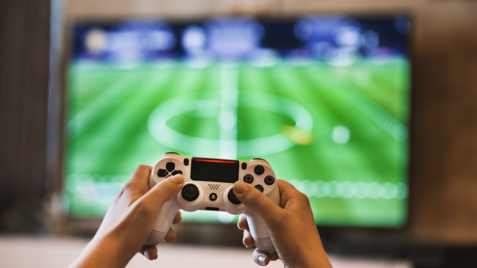 Hands holding a gaming controller in front of a television displaying a sporting event.