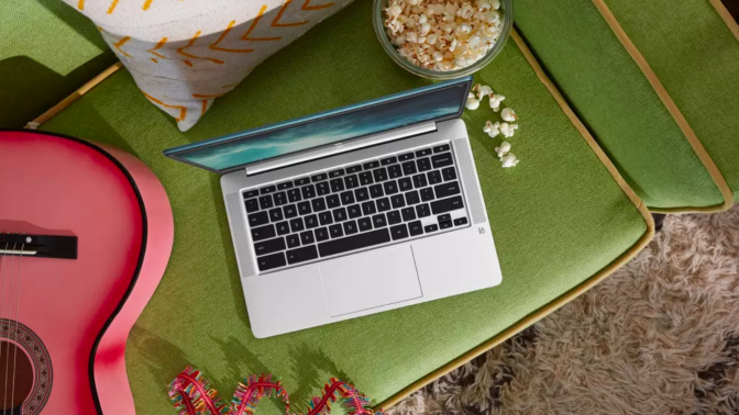 a top-down view of an hp chromebook sitting on an olive green couch next to a pink guitar and a spilled bowl of popcorn